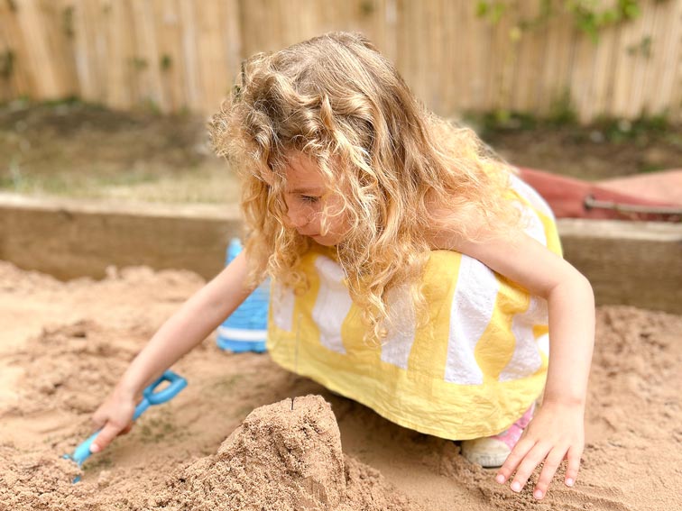 a young girl with long blonde curly hair and a yellow dress playing in a sand pit