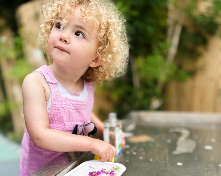 a young girl with blonde curly hair and pink overalls playing with water and flowers and looking up to the left of the camera