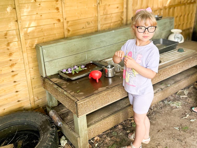 a young blonde girl with glasses and a fringe playing in an outdoor kitchen area, looking at something in the distance