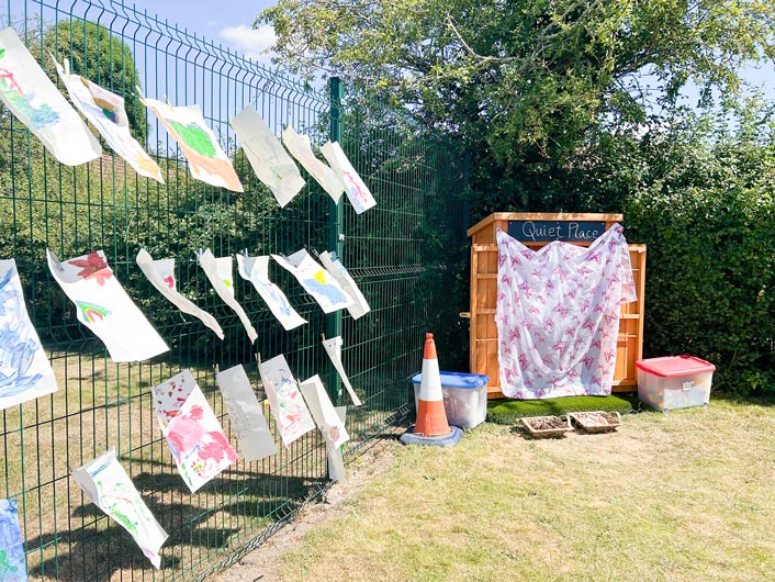 children's paintings drying outside in the sun on a green fence with a shed next to them that says 'quiet place'