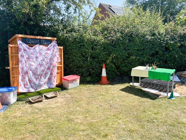 a garden space in the sun with a shed to the left that says 'quiet place' and a table to the right with paints set up