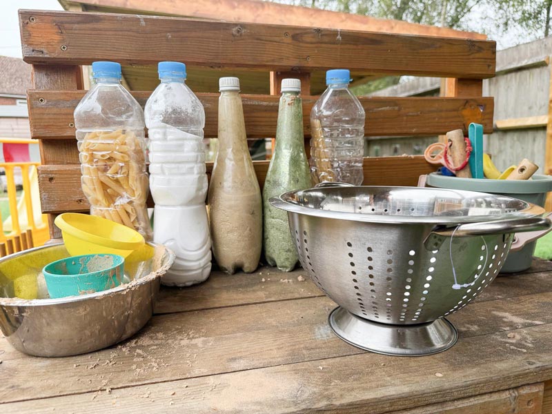 a wooden table set up with children's play activities including bottles full of sand, a colander, and sand toys
