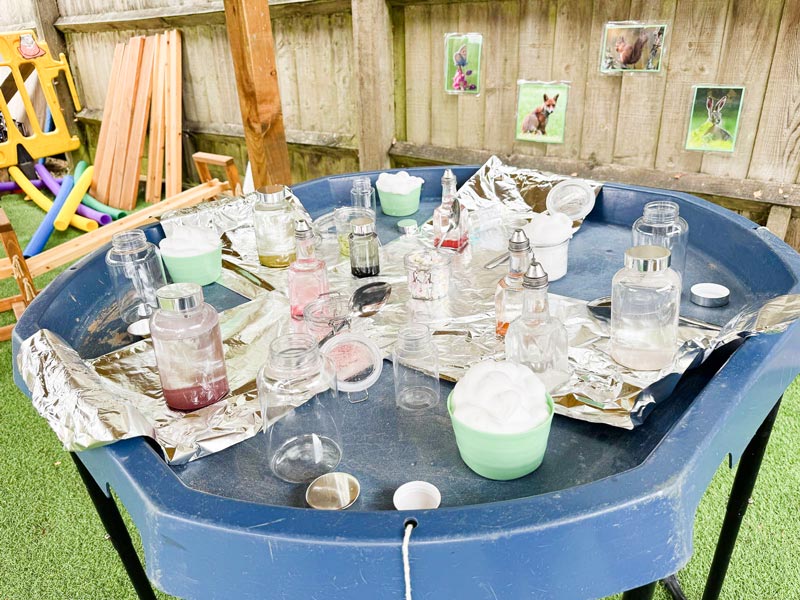 a play table with glass jars and bottles with different colour liquids, aluminium foil, bowls of cotton balls and spoons