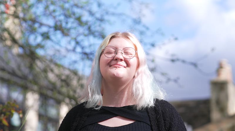 a young women with short blonde hair and glasses standing in the sunshine smiling with her eyes closed
